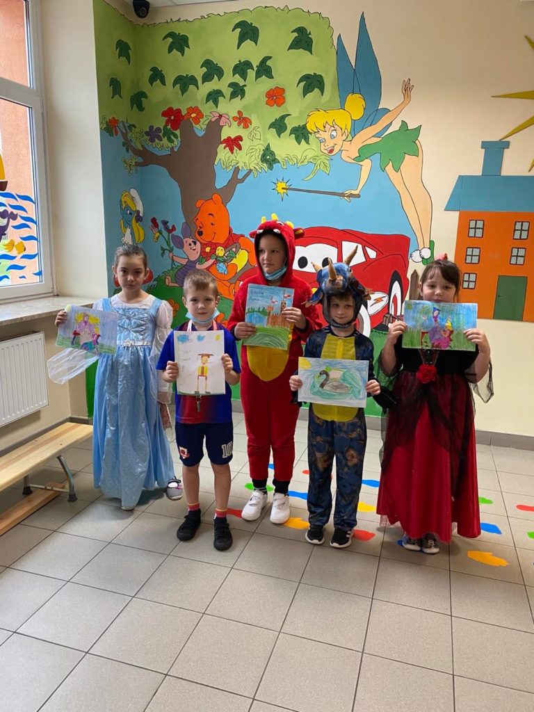 Grupa uczniów z klasy drugiej przebrani za wróżki, czerwony kapturek, księżniczkę  stoją a w ręku trzymają namalowane postaci z ulubionych bajek. 