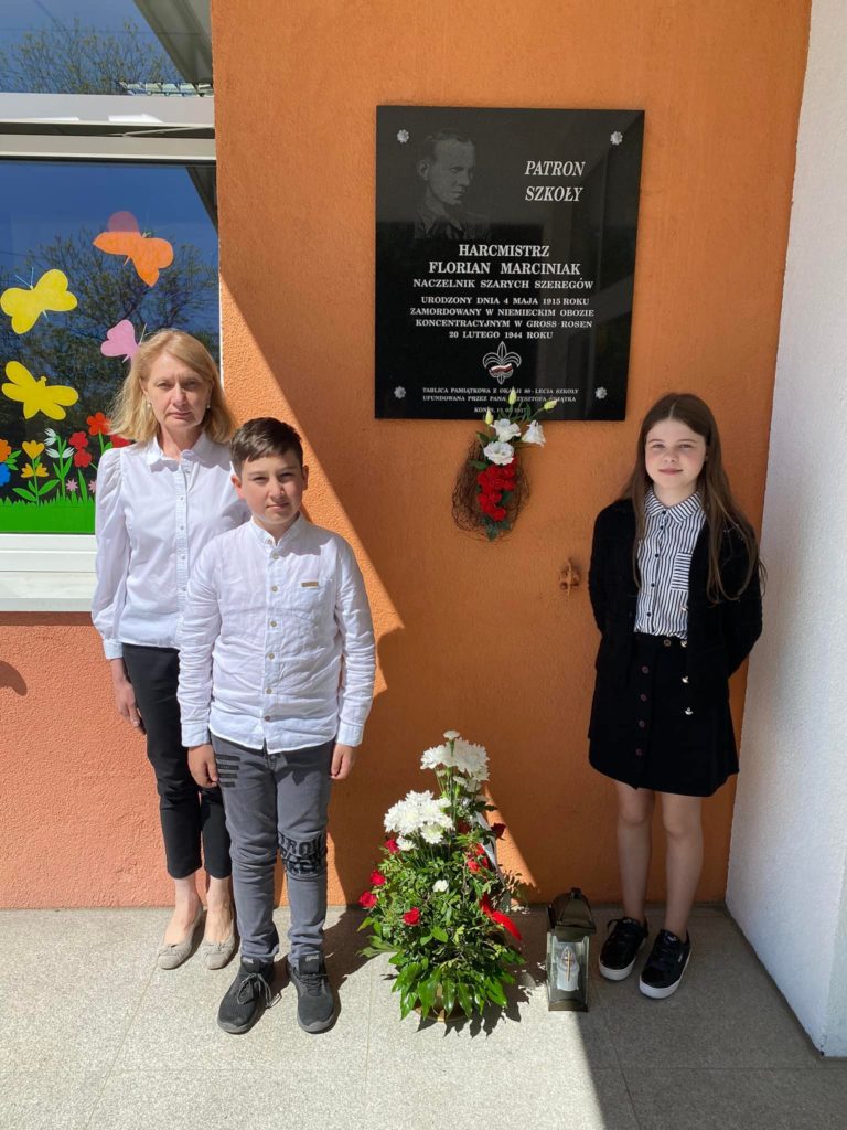 Na fotografia jest dwójka uczniów wraz z panią Agnieszką Czechowską pod tablicą patrona szkoły. 