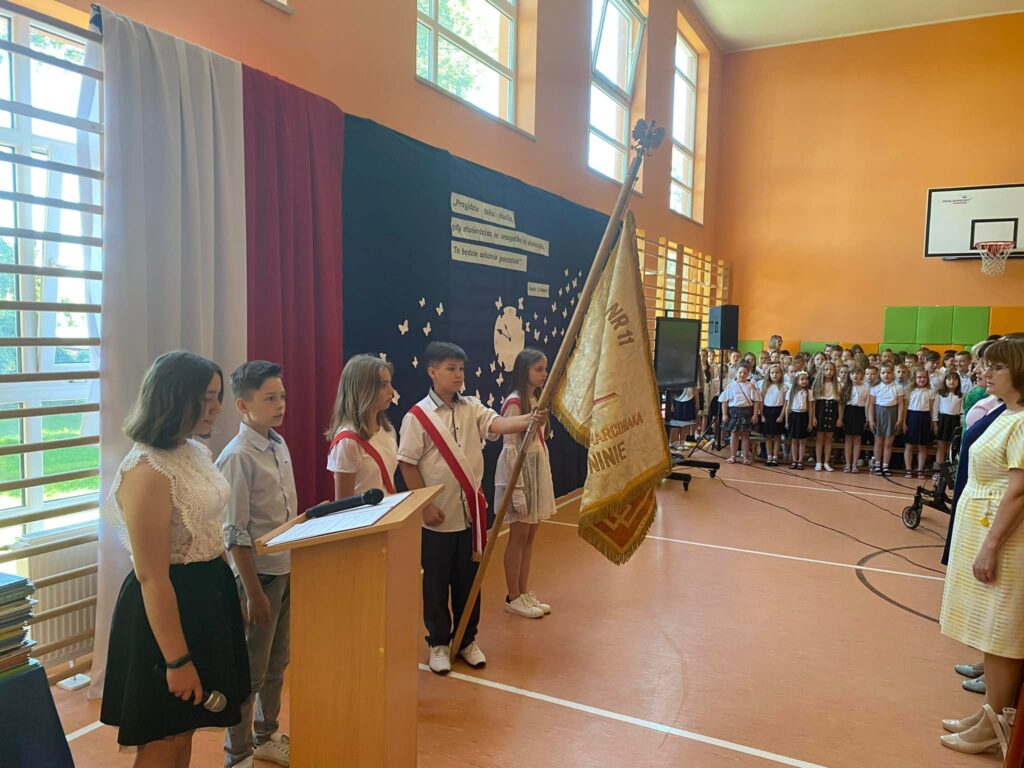 Fotografia przedstawia poczet szandarowy szkoły podczas śpiewania hymnu państwowego. 