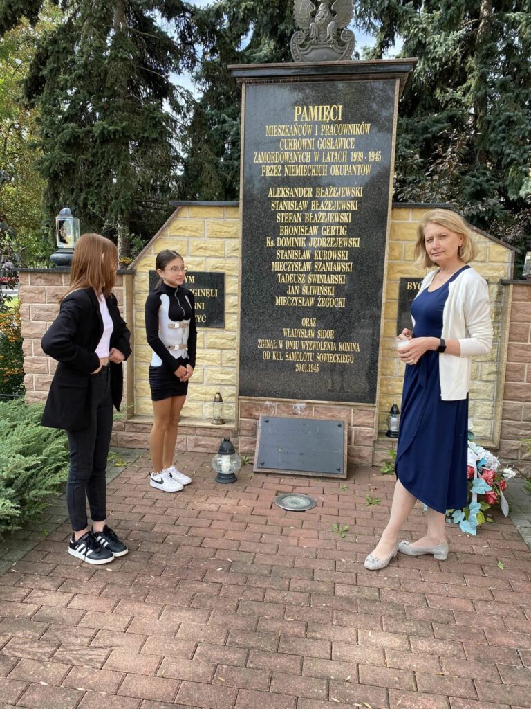 Fotografia przedstawia dwie uczennice z klasy ósmej b wraz z panią Agnieszką Czechowską pod pomnikiem upamiętniającym ofiary II wojny światowej. 
