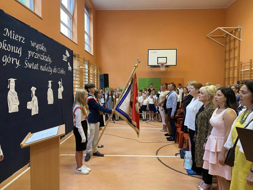 Fotografia przedstawia społeczność szkolną podczas śpiewania hymnu państwowego.