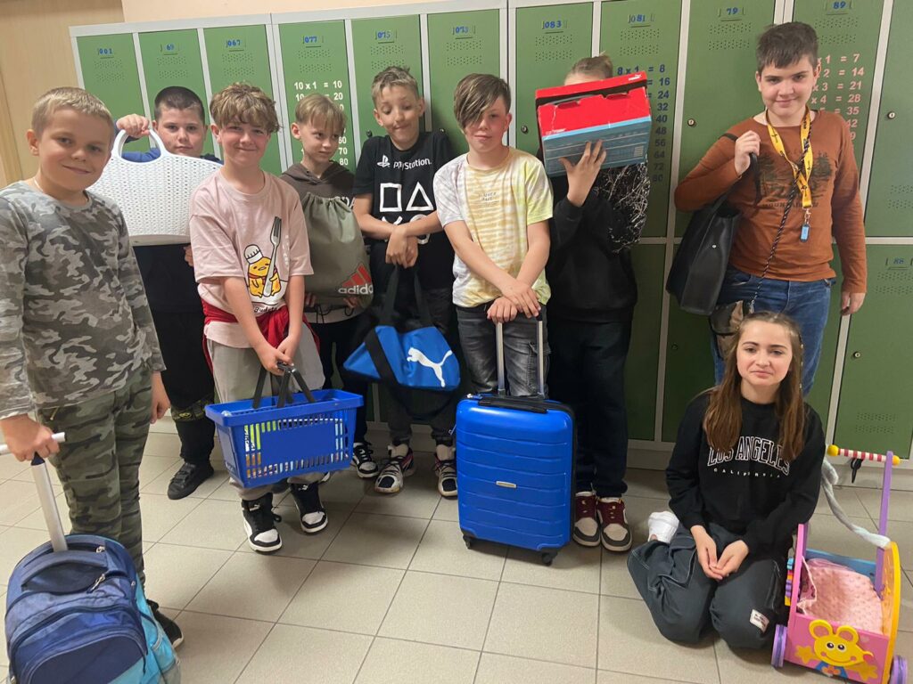 Fotografia przedstawia uczniów klasy piątek trzymających koszyki, walizki, kartony zamiast plecaków.