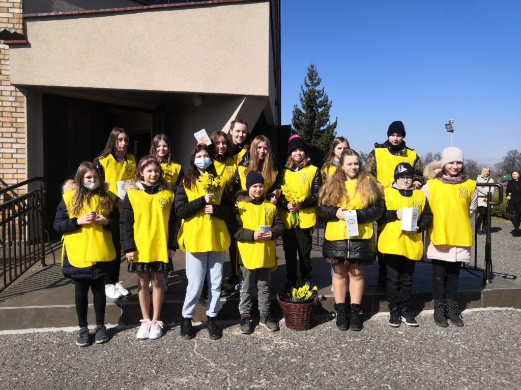 Fotografia przedstawia wolontariuszy z naszej szkoły pod kościołam. Uczniowie ubrani są w żółte kamizelki.