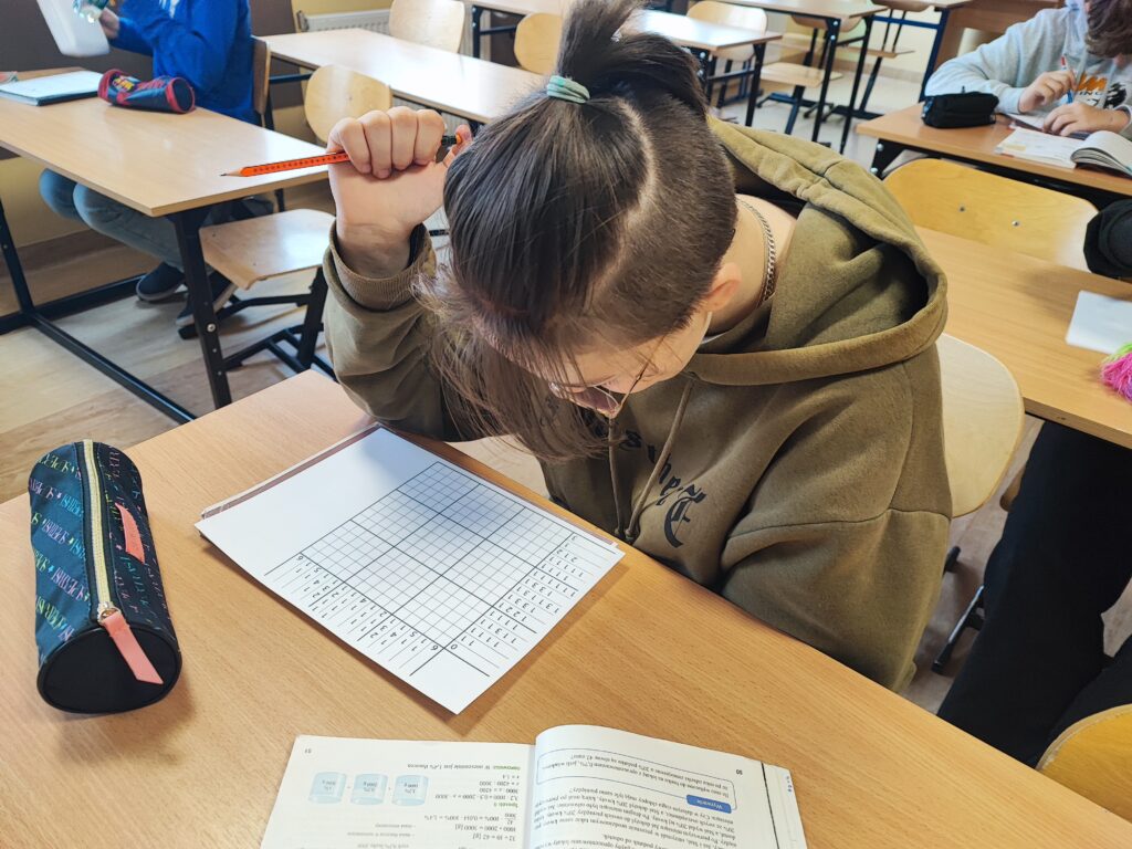 Fotografia przedstawia uczennice klasy siódmej rozwiązująca zagadkę logiczną na kartce. 