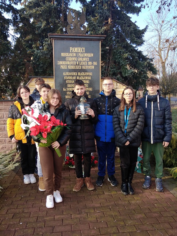 Fotografia przedstawia uczniów klasy siódmej trzymających kwiaty pod pomnikiem.