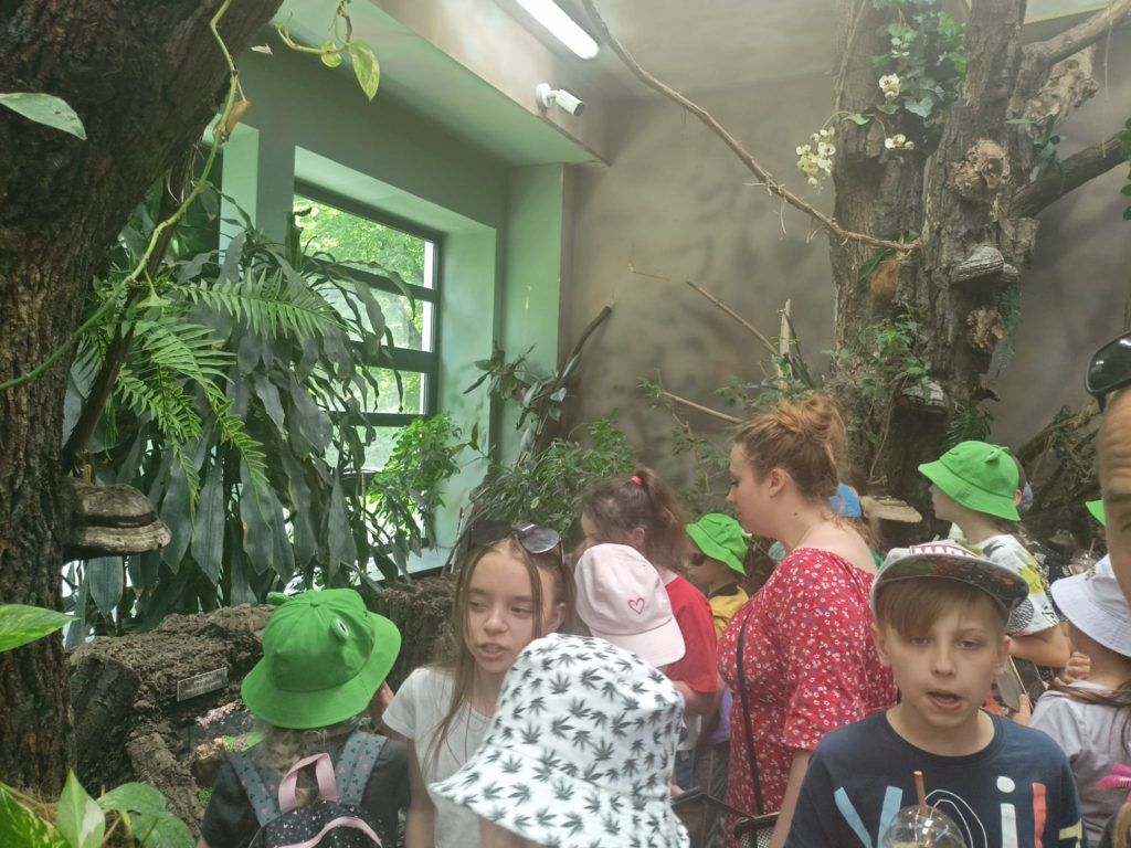 Zdjęcie przedstawia grupę uczniów stojący przy bujnej roślinności na terenie zoo. 