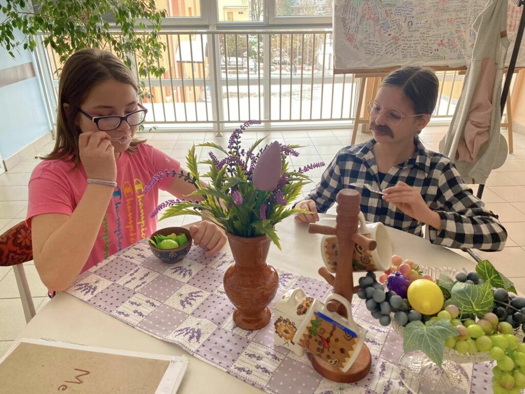 dwie uczennice siedzą przy stoliku nakrytym owocami, talerzami. Odgrywają scenkę w restauracji