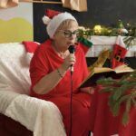 pani w czapce Mikołaja siedzi na czerwonym fotelu trzyma mikrofon i czyta dzieciom bajkę w tle kominek i dekoracja świąteczna