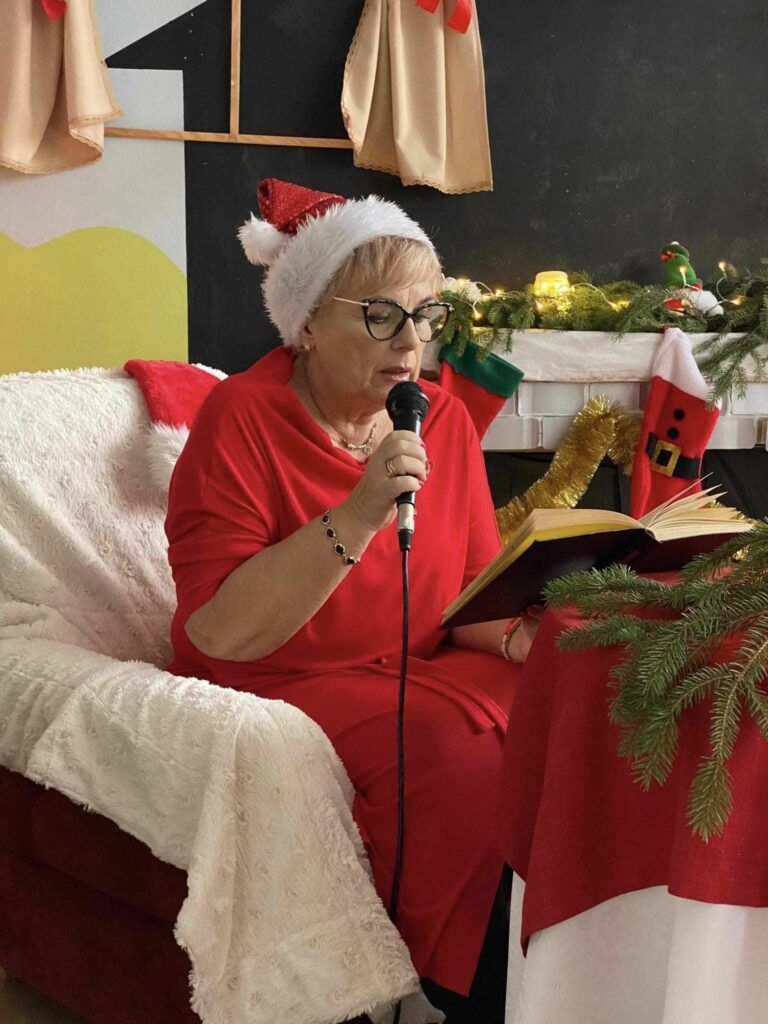 pani w czapce Mikołaja siedzi na czerwonym fotelu trzyma mikrofon i czyta dzieciom bajkę w tle kominek i dekoracja świąteczna