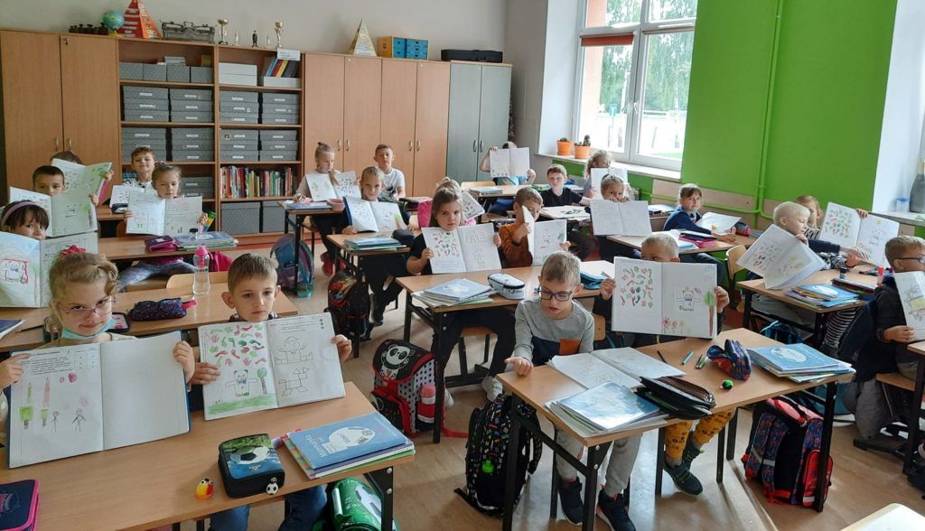Fotografia przedstawiająca uczniów klasy pierwszej siedzących przy ławkach i pokazujących swoje zeszyty.